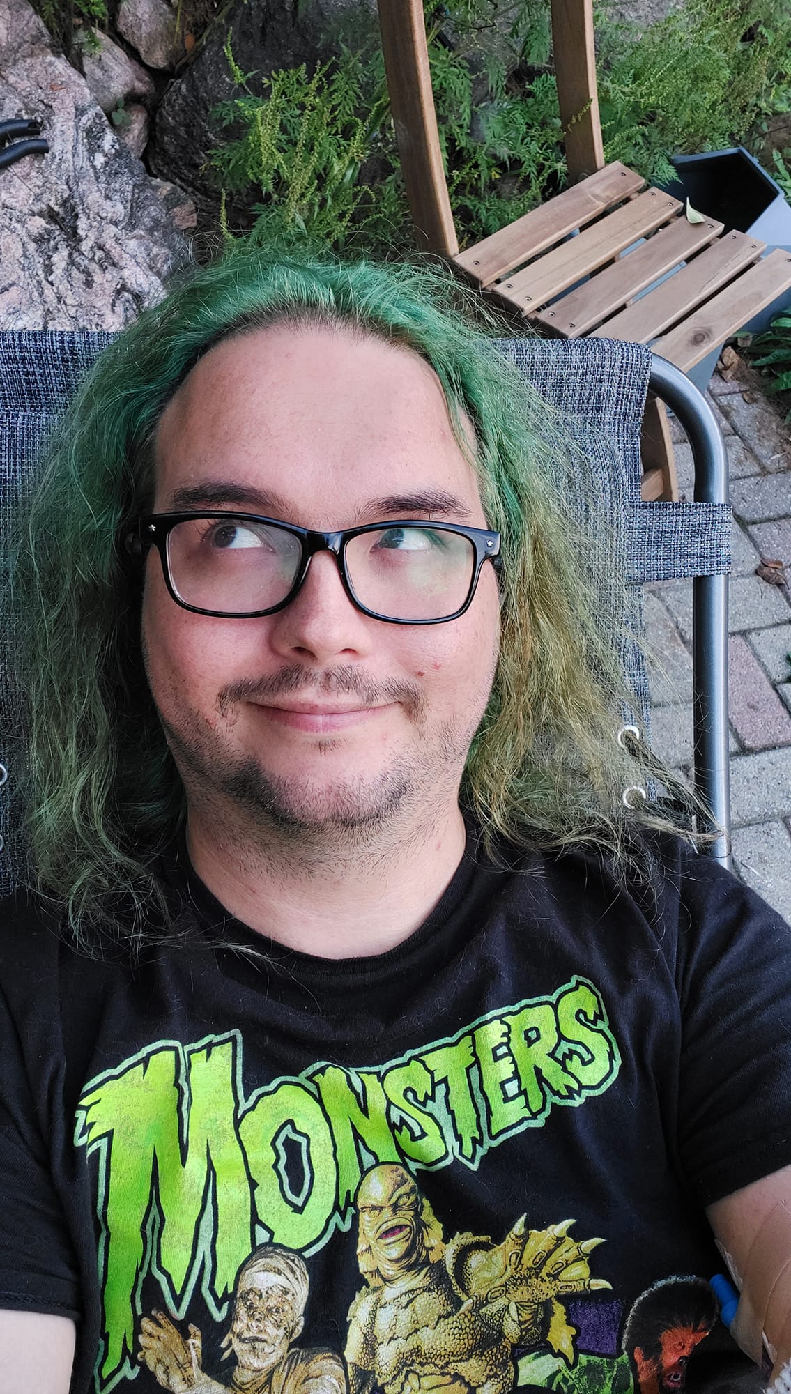 A man with long green hair and no beard sits lays in a folding chair on a patio, Photo 1